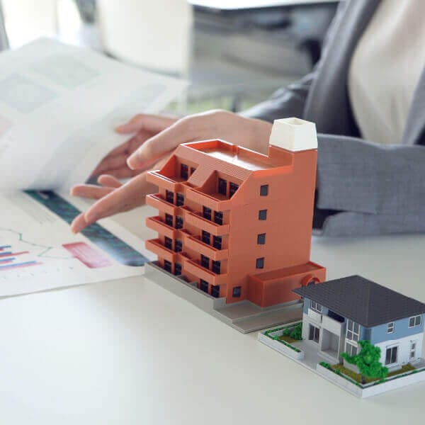 机の上にマンションと戸建て住宅の建物模型が置かれ、不動産資料を確認する人物の手元