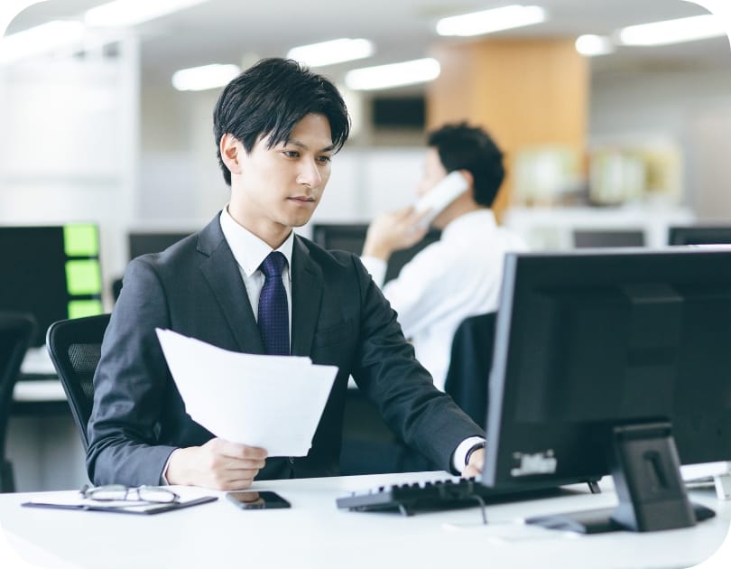 スーツ姿のビジネスマンがデスクで書類を手にしながらパソコンを操作している