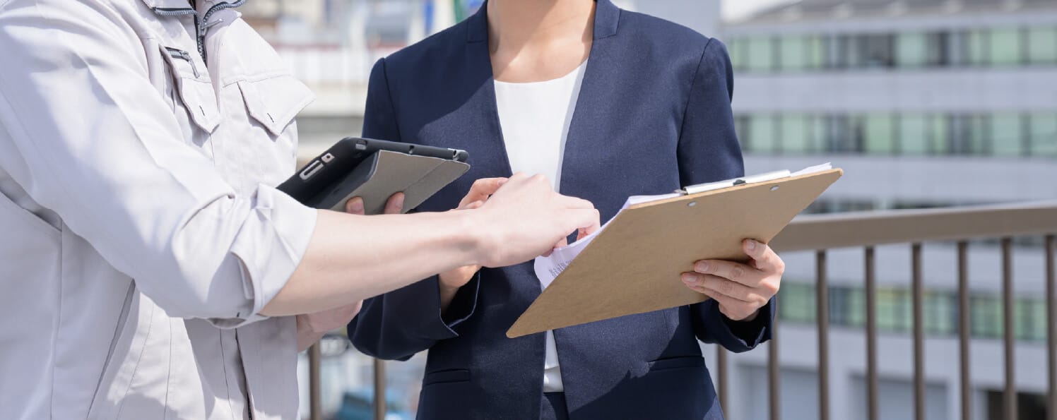 作業服の人物とスーツ姿の人物がクリップボードを見ながら屋外で打ち合わせ中