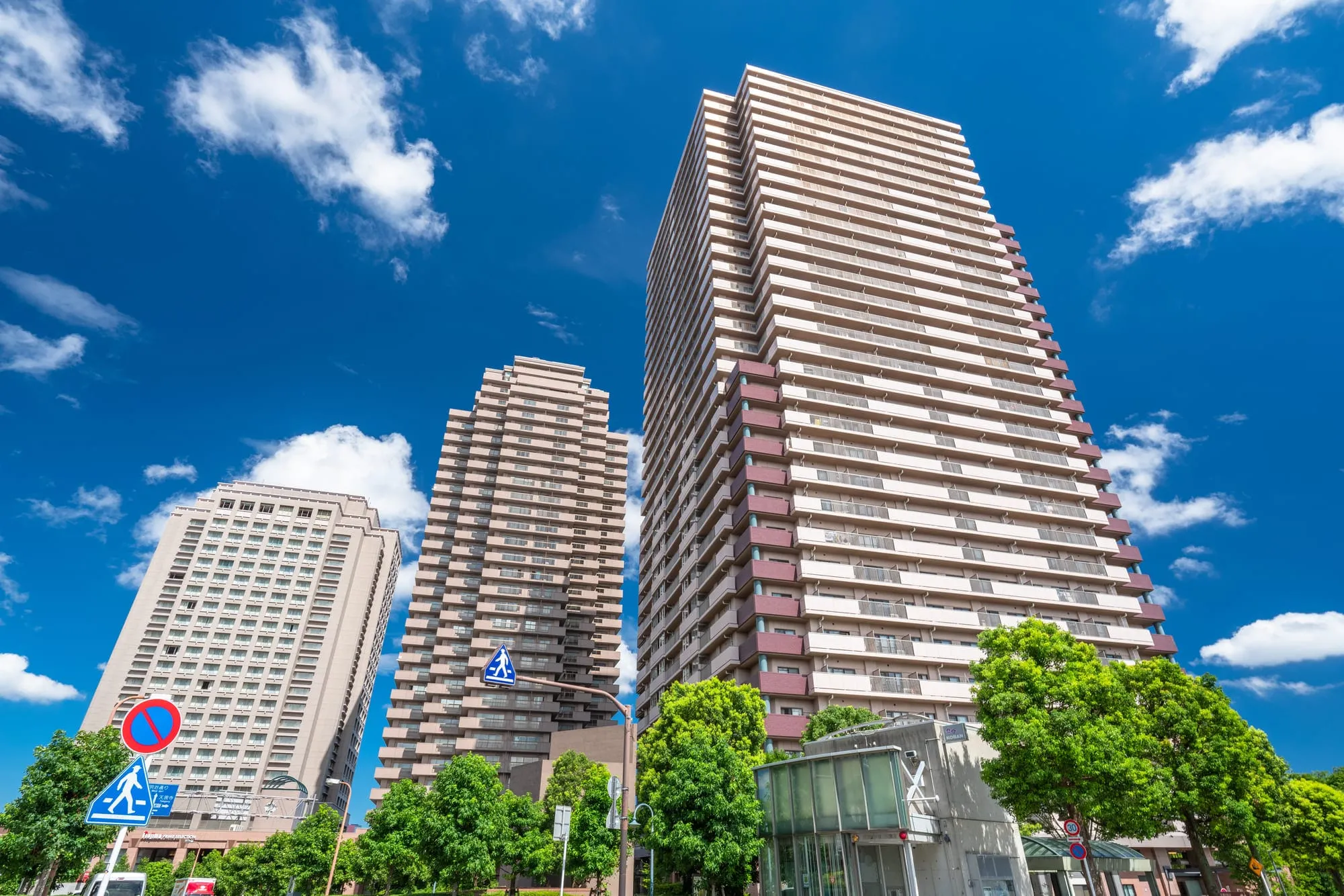 青空のもとにそびえるタワーマンション群を見上げた構図で、手前には歩道と標識が写っている